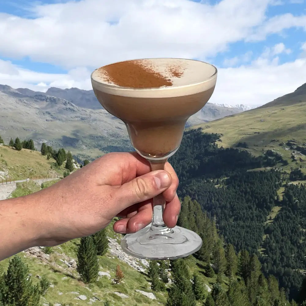 Koktajl Chaitini z chai masala w kieliszku, trzymany na tle górskiego krajobrazu – napój na bazie herbaty z przyprawami, w wersji lifestyle.