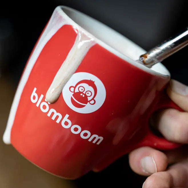 Czerwona filiżanka z logo "blömboom" i grafiką małpy, z której spływa mleczna piana, trzymana w dłoni baristy.