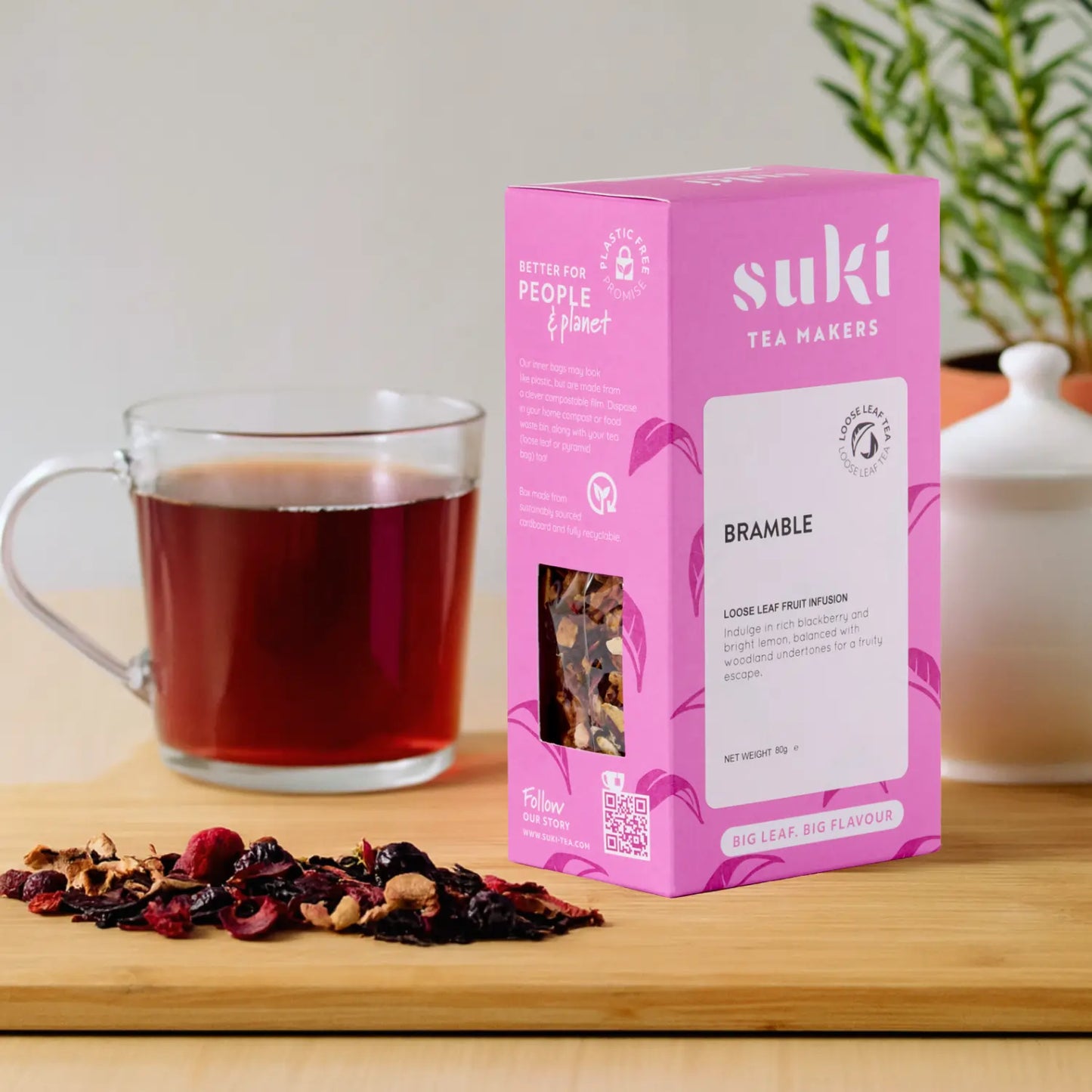 Herbata owocowa SUKI Tea Bramble – różowe opakowanie 80 g z suszonymi owocami i szklanką zaparzonego naparu na drewnianym blacie.
