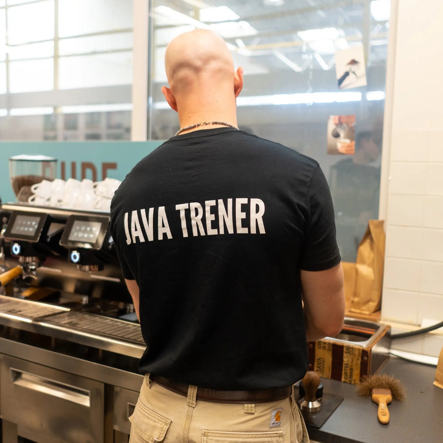 Trener JAVA Coffee Roasters podczas szkolenia baristycznego przy ekspresie do kawy, widok od tyłu z napisem „JAVA TRENER” na koszulce.