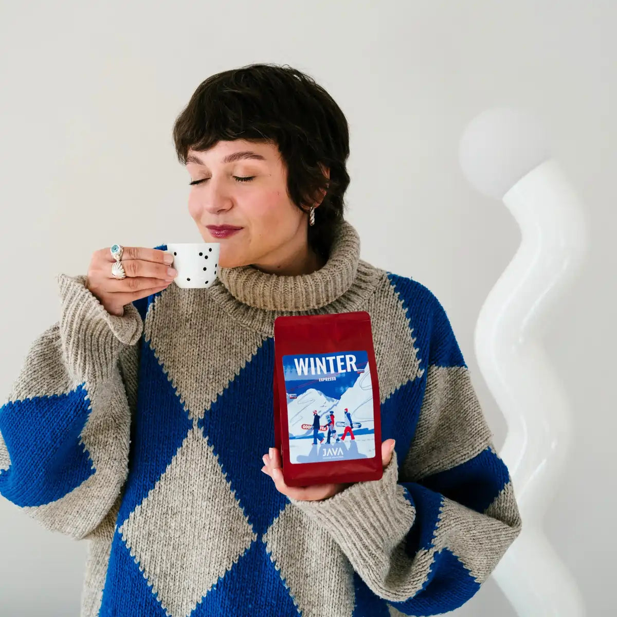 Osoba trzyma czerwone opakowanie kawy JAVA Coffee Winter Espresso i pije kawę z małej filiżanki, w jasnym domowym wnętrzu.