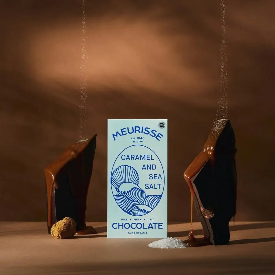 Tabliczka belgijskiej mlecznej czekolady Meurisse Caramel & Sea Salt w niebieskim opakowaniu, otoczona karmelem i kryształkami soli morskiej na brązowym tle.