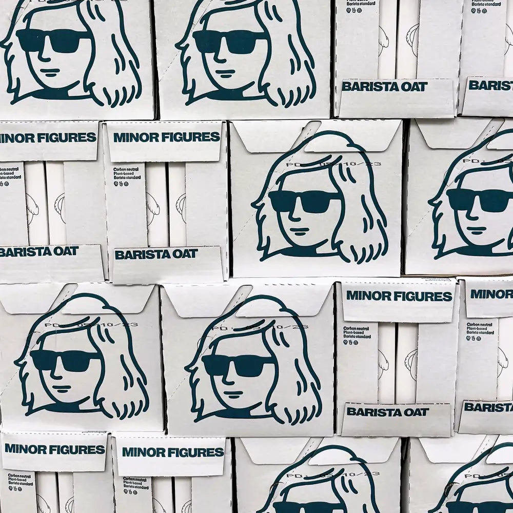 Zbliżenie na ułożone kartony z napojem owsianym Minor Figures Barista Oat – białe pudełka z grafiką postaci w okularach przeciwsłonecznych i napisem „Carbon neutral, Plant-based, Barista standard”.