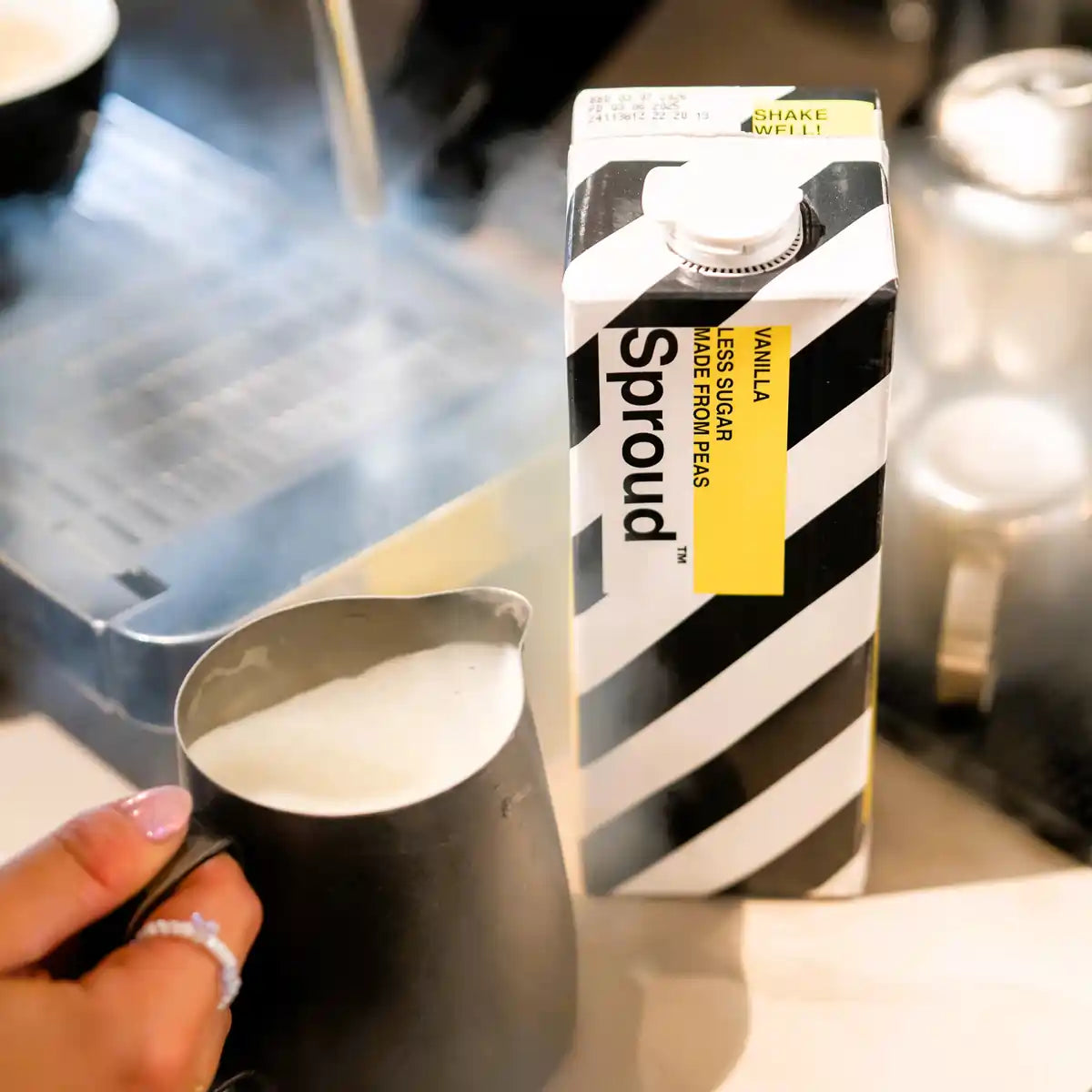 Karton napoju roślinnego Sproud Vanilla 1L obok dzbanka z pienionym mlekiem – wegański napój z grochu o smaku waniliowym, idealny do kawy i deserów.