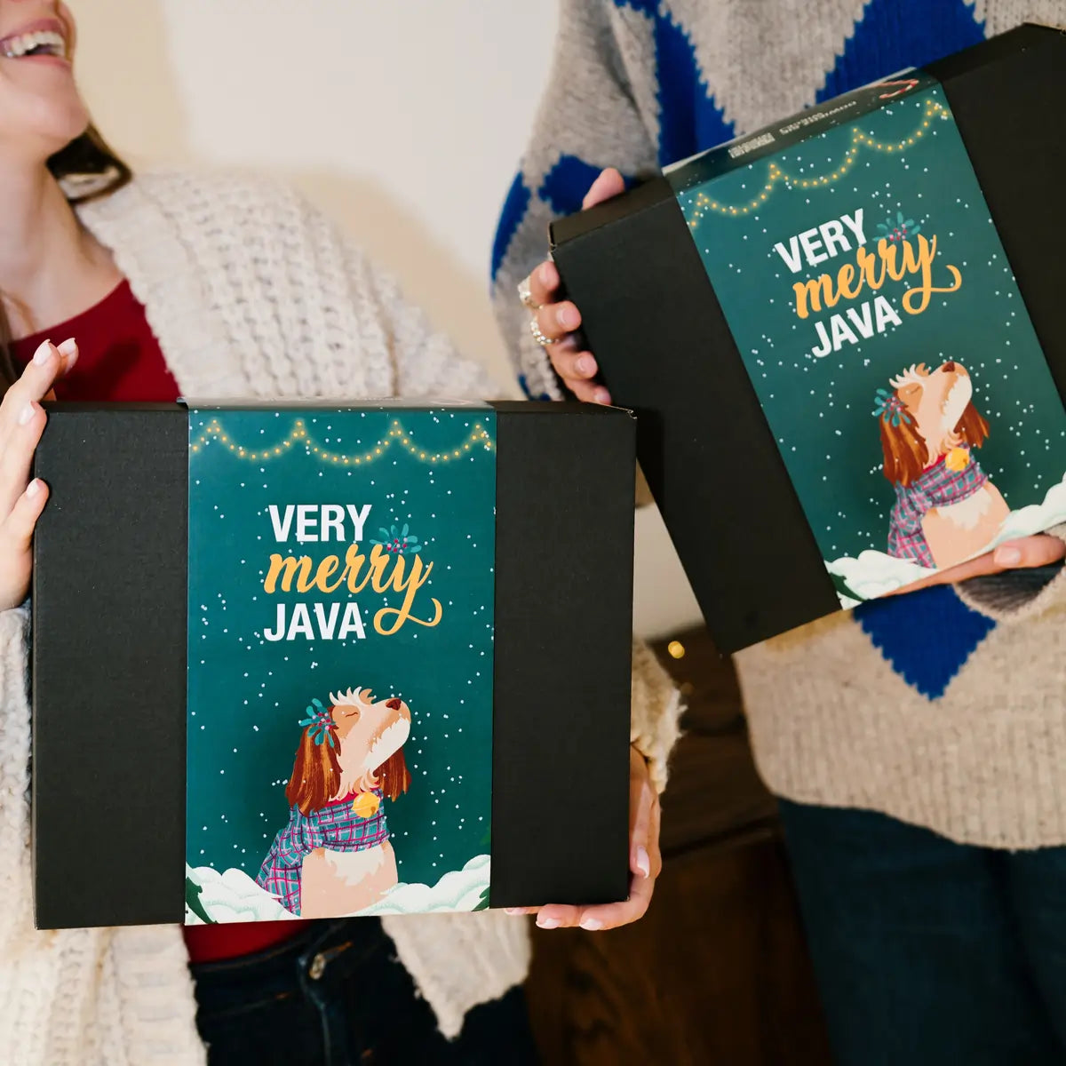Świąteczne pudełko prezentowe Very Merry JAVA w czarnym opakowaniu z zieloną kartonową owijką, trzymane przez dwie osoby w zimowych swetrach.