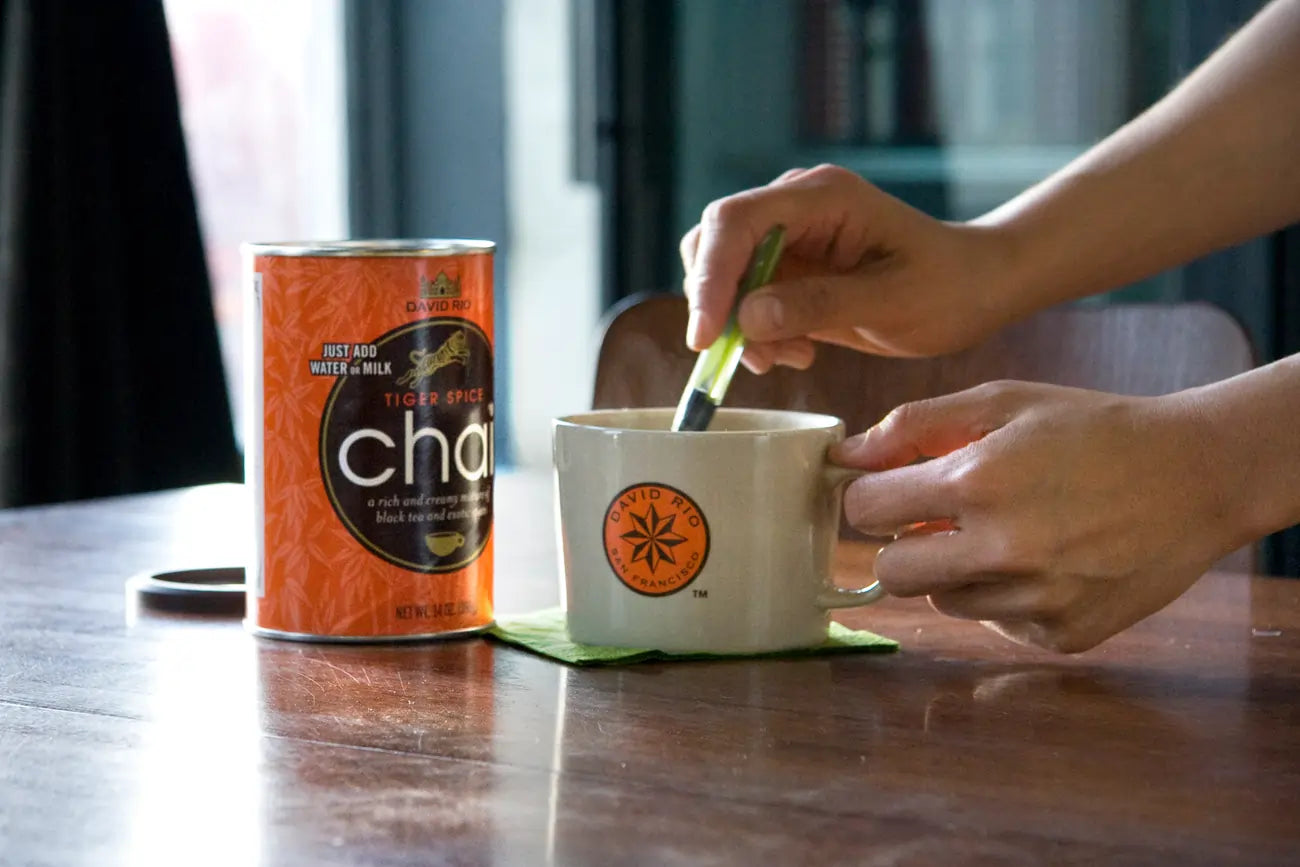 Dłonie przygotowujące chai latte w kremowym kubku na drewnianym stole, obok znajduje się pomarańczowa puszka Chai.