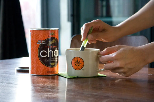 Dłonie przygotowujące chai latte w kremowym kubku na drewnianym stole, obok znajduje się pomarańczowa puszka Chai.