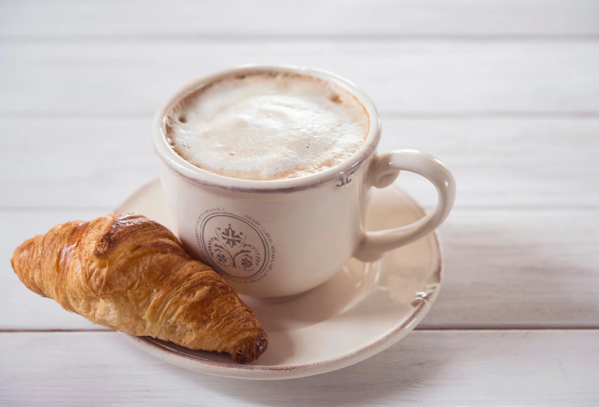 Cappucino z gęstą i puszystą pianką na spodeczku, na którym jest także croissont – idealny zestaw na śniadanie, ustawiony na jasnym, drewnianym stole.