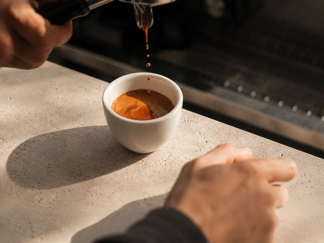 Espresso lungo being poured from an espresso machine into a white cup, with the barista's hands visible at the counter.