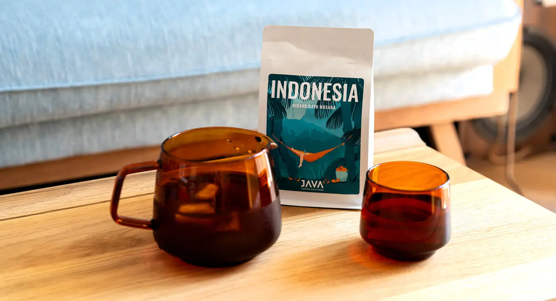 Opakowanie kawy Indonesia Ribang Gayo Musara od JAVA Coffee ustawione na drewnianym stole obok dwóch bursztynowych filiżanek z kawą - blog o kawie JAVA Coffee
