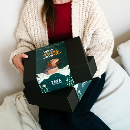 Świąteczne pudełko prezentowe JAVA Coffee Roasters „Very Merry Java” trzymane na kolanach, czarne pudełko z zimową grafiką i psem w szaliku, ujęcie lifestyle w domowym wnętrzu.