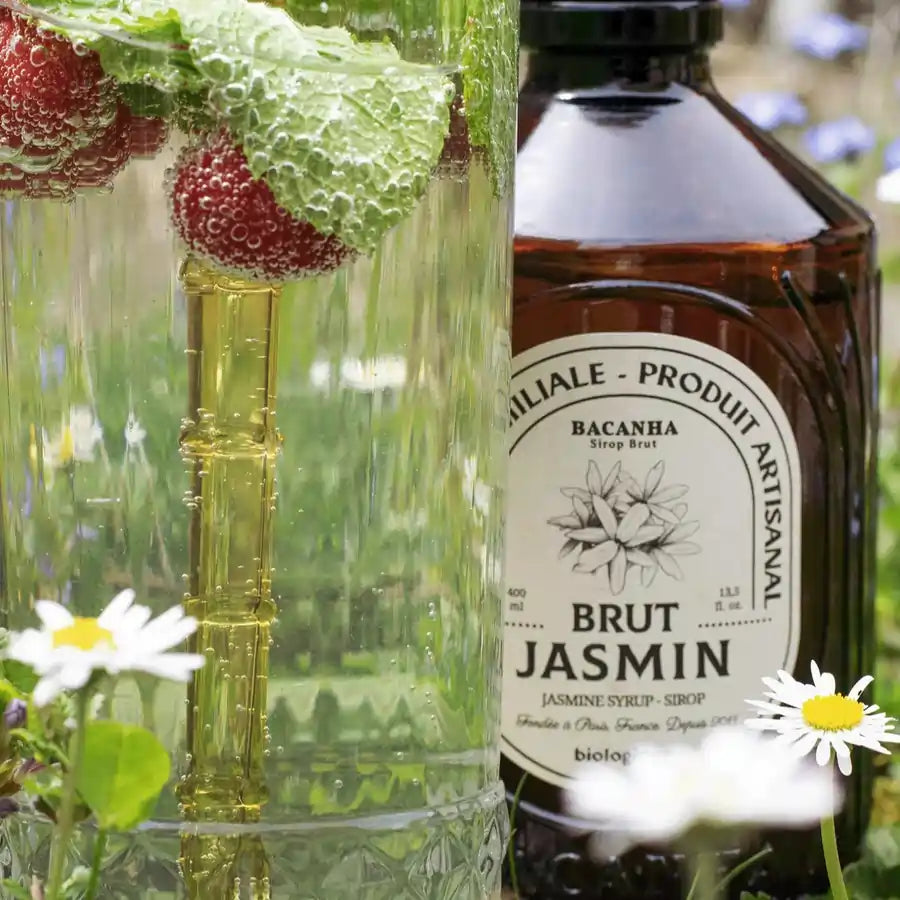 Syrop Bacanha o smaku jaśminowym obok szklanki z napojem, malinami i listkami mięty wśród kwiatów – letnia, kwiatowa kompozycja.