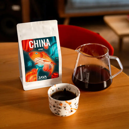 Kawa specialty China Fuyan od JAVA Coffee Roasters – opakowanie kawy, szklany dzbanek z kawą przelewową oraz filiżanka espresso na drewnianym stole, zdjęcie lifestyle