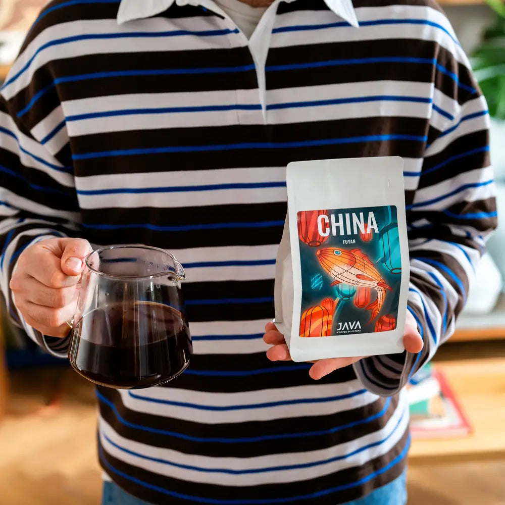 Kawa specialty China Fuyan od JAVA Coffee Roasters – zaparzona kawa przelewowa trzymana w szklanym dzbanku oraz opakowanie kawy ziarnistej w dłoni