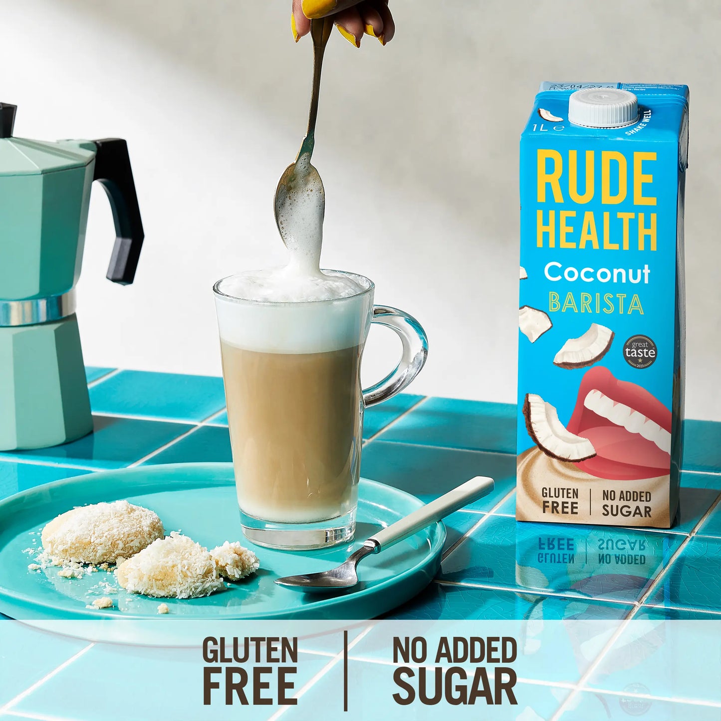 Szklanka kokosowego latte przygotowanego na mleku Rude Health Coconut Barista, obok kartonika napoju i kokosowych ciasteczek