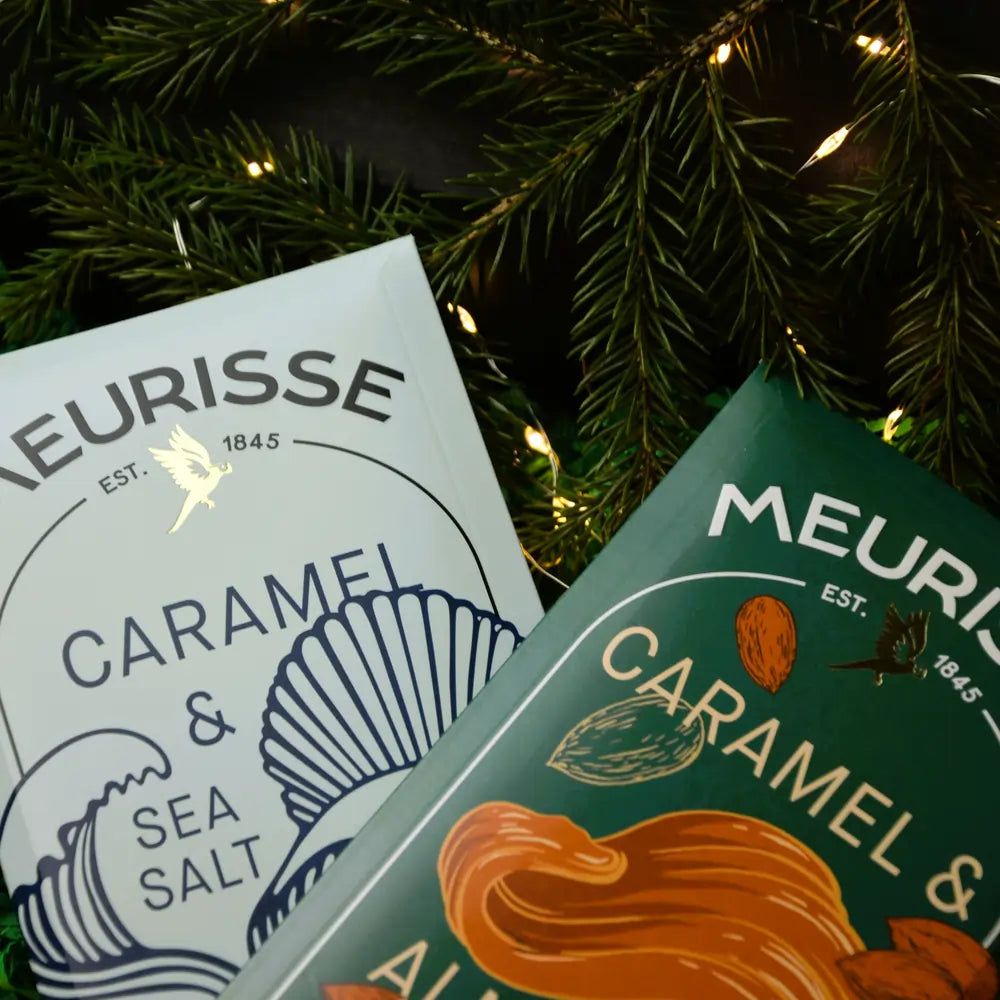 Świąteczne zbliżenie na tabliczki czekolady Meurisse Caramel & Sea Salt oraz Caramel & Almonds w otoczeniu gałązek świerku i lampek.