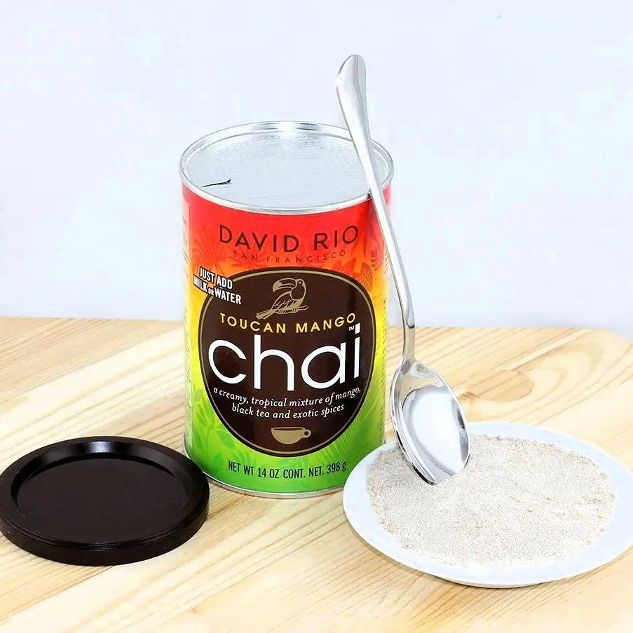 David Rio Toucan Mango Chai 398 g – napój chai w proszku z odsłoniętą puszką, łyżeczką i porcją proszku na talerzyku w ujęciu lifestyle