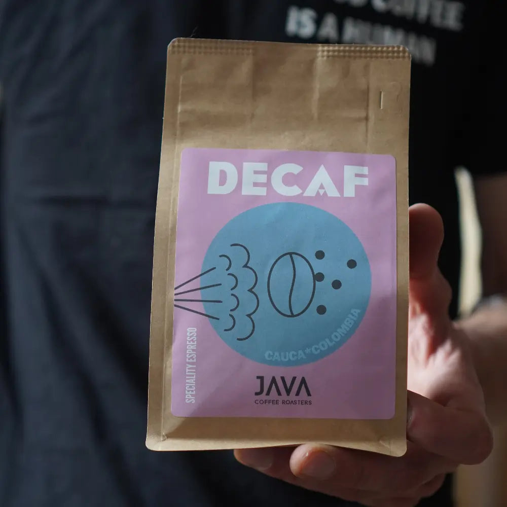 Brązowa torebka kawy Java Coffee Roasters z etykietą „DECAF” w różowo-niebieskich kolorach, trzymana w dłoni osoby w czarnej koszulce z napisem „IS A HUMAN”. Na etykiecie widoczne są ilustracje ziaren kawy i bąbelków oraz napis „CAUCA • COLOMBIA”