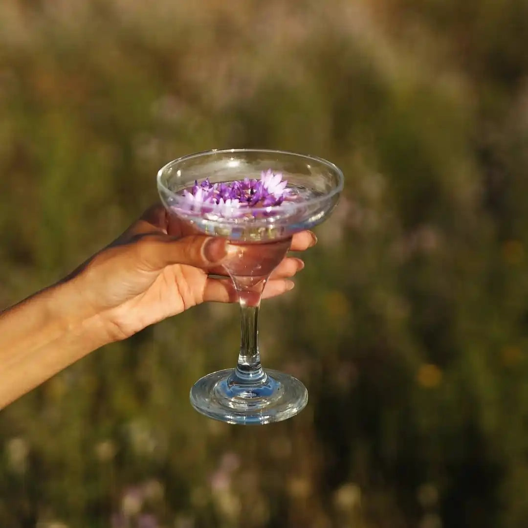 Osoba trzyma kieliszek z przezroczystym napojem ozdobionym fioletowymi płatkami kwiatów na tle zieleni natury