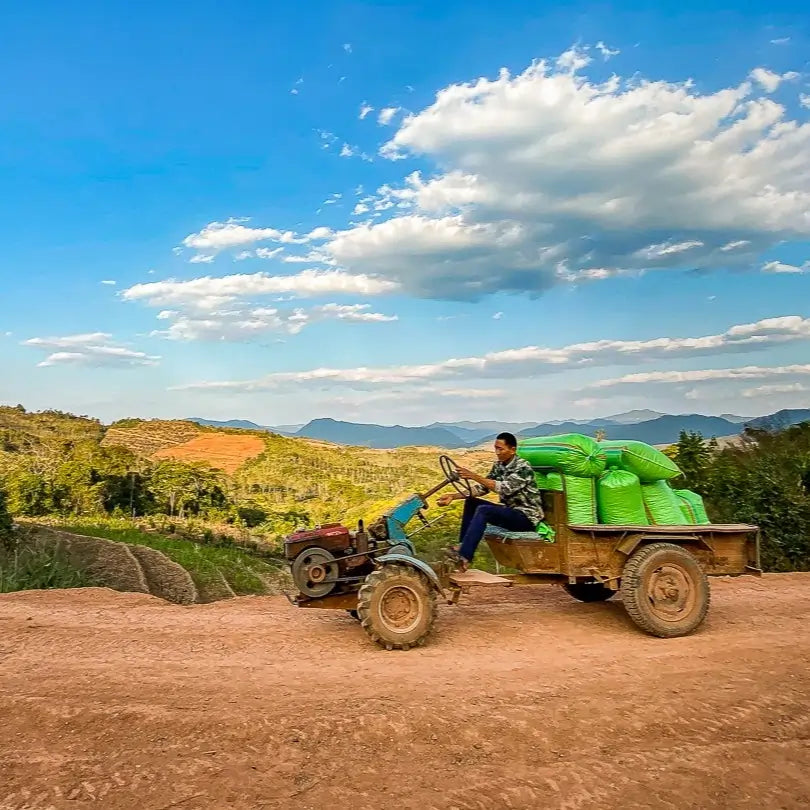 Rolnik jadący małym traktorem z przyczepą pełną zielonych worków po polnej drodze w górzystym krajobrazie pod błękitnym niebem.