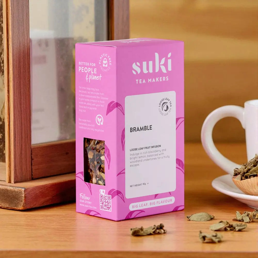 Opakowanie herbaty owocowej SUKI Tea Bramble 80 g – różowe pudełko na drewnianym blacie, obok filiżanki i suszonych liści.