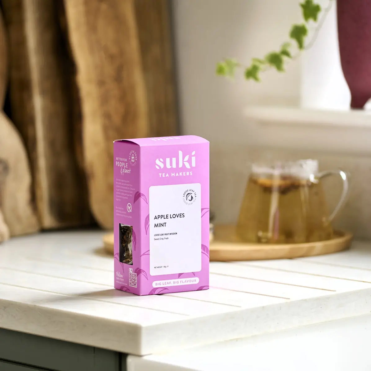 Herbata Suki Apple Loves Mint w różowym opakowaniu stojąca na kuchennym blacie, w tle dzbanek z zaparzoną herbatą i drewniana taca.