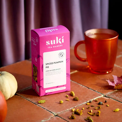 Herbata Suki Spiced Pumpkin Pie w różowym opakowaniu na stole z filiżanką zaparzonej herbaty w tle, jesienna kompozycja z dynią i przyprawami.