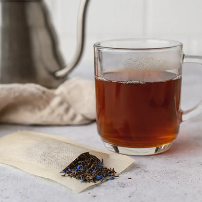 Szklanka gorącej czarnej herbaty obok saszetki z herbatą liściastą i płatkami bławatka na jasnym blacie.