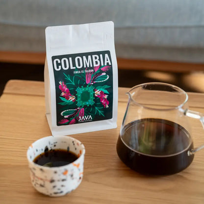 Opakowanie kawy specialty JAVA Coffee na drewnianym stole. Etykieta z napisem 'Colombia' i kolorową grafiką inspirowaną naturą. 