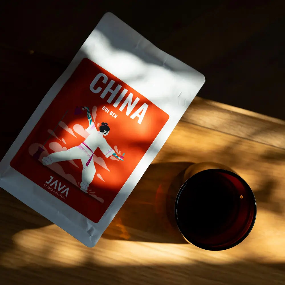 Paczka kawy speciality JAVA Coffee Roasters China Gui Ben na drewnianym stole w promieniach słońca obok filiżanki zaparzonej kawy.