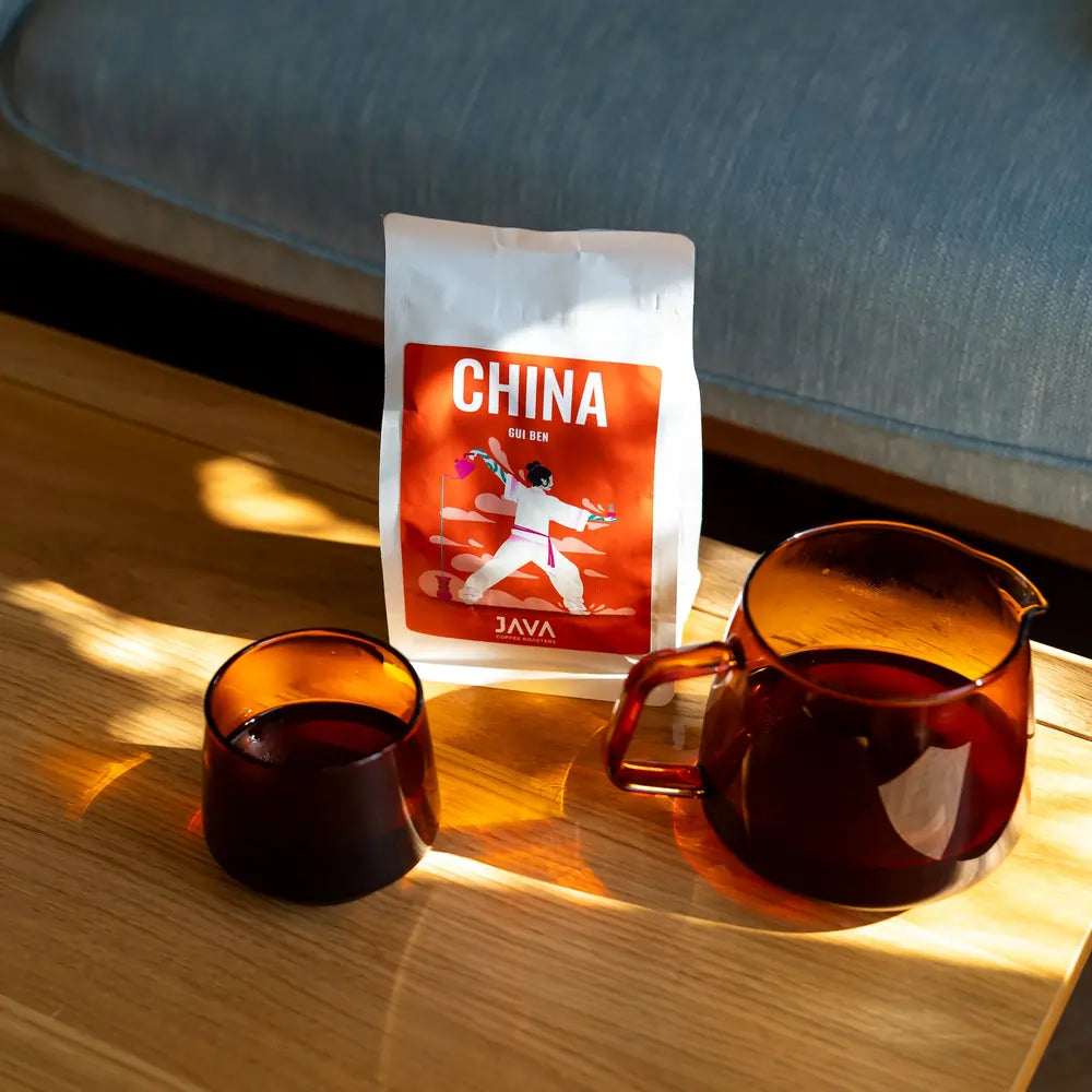 Kawa JAVA Coffee China Gui Ben stojąca na drewnianym stole obok dzbanka i filiżanki z zaparzoną kawą w ciepłym świetle słońca.