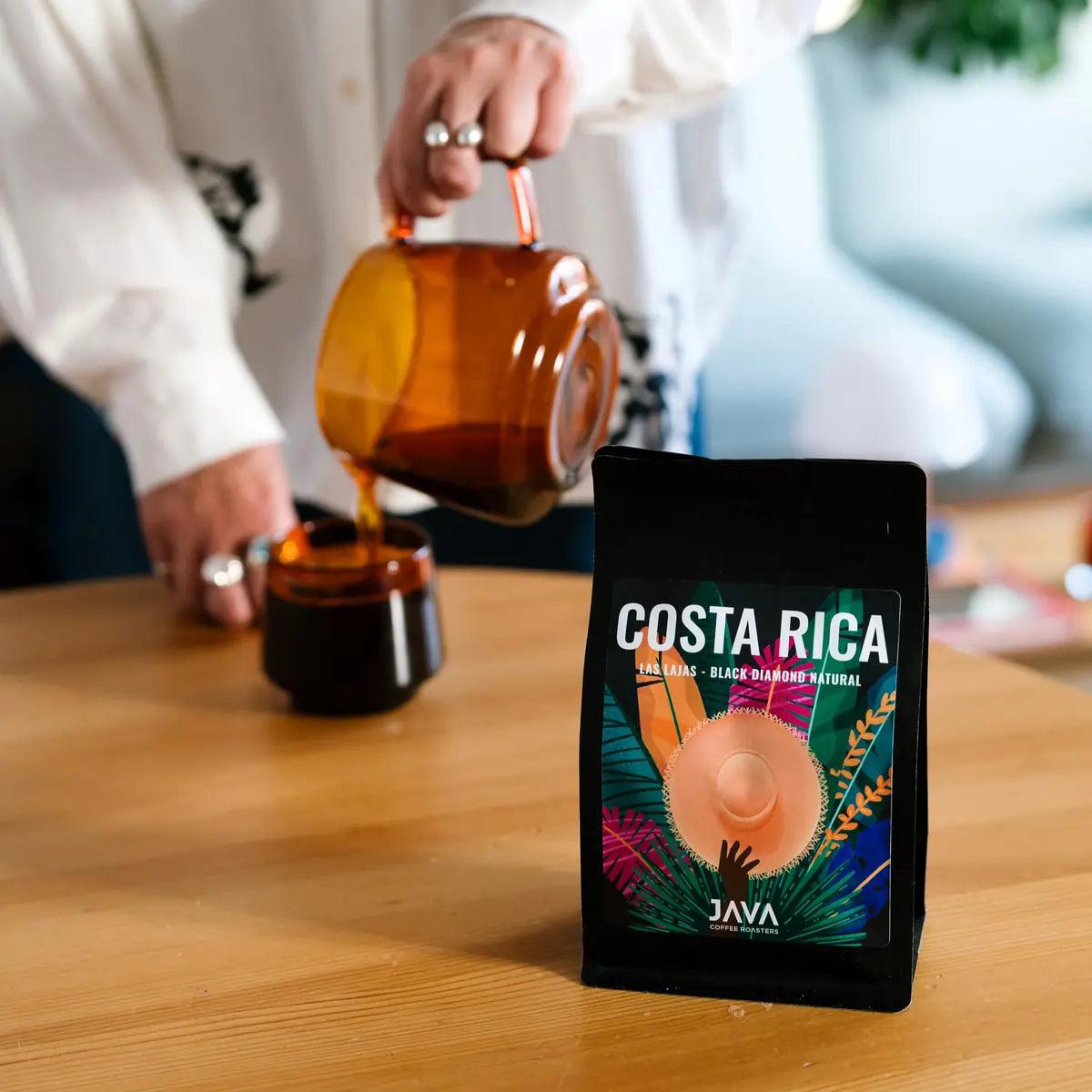 Kawa specialty JAVA Coffee Costa Rica Las Lajas Black Diamond Natural na stole, w tle osoba nalewająca kawę z dzbanka do bursztynowej filiżanki