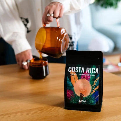 Kawa specialty JAVA Coffee Costa Rica Las Lajas Black Diamond Natural na stole, w tle osoba nalewająca kawę z dzbanka do bursztynowej filiżanki
