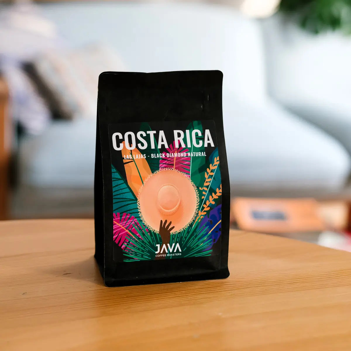 Kawa specialty JAVA Coffee Costa Rica Las Lajas Black Diamond Natural w czarnym opakowaniu stojąca na drewnianym stole, w tle jasne wnętrze z roślinami