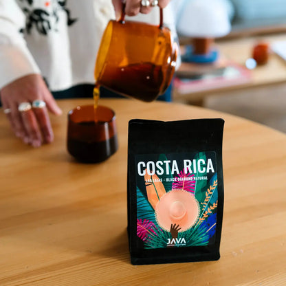 Kawa specialty JAVA Coffee Costa Rica Las Lajas Black Diamond Natural w czarnym opakowaniu na stole, w tle osoba nalewająca kawę do filiżanki