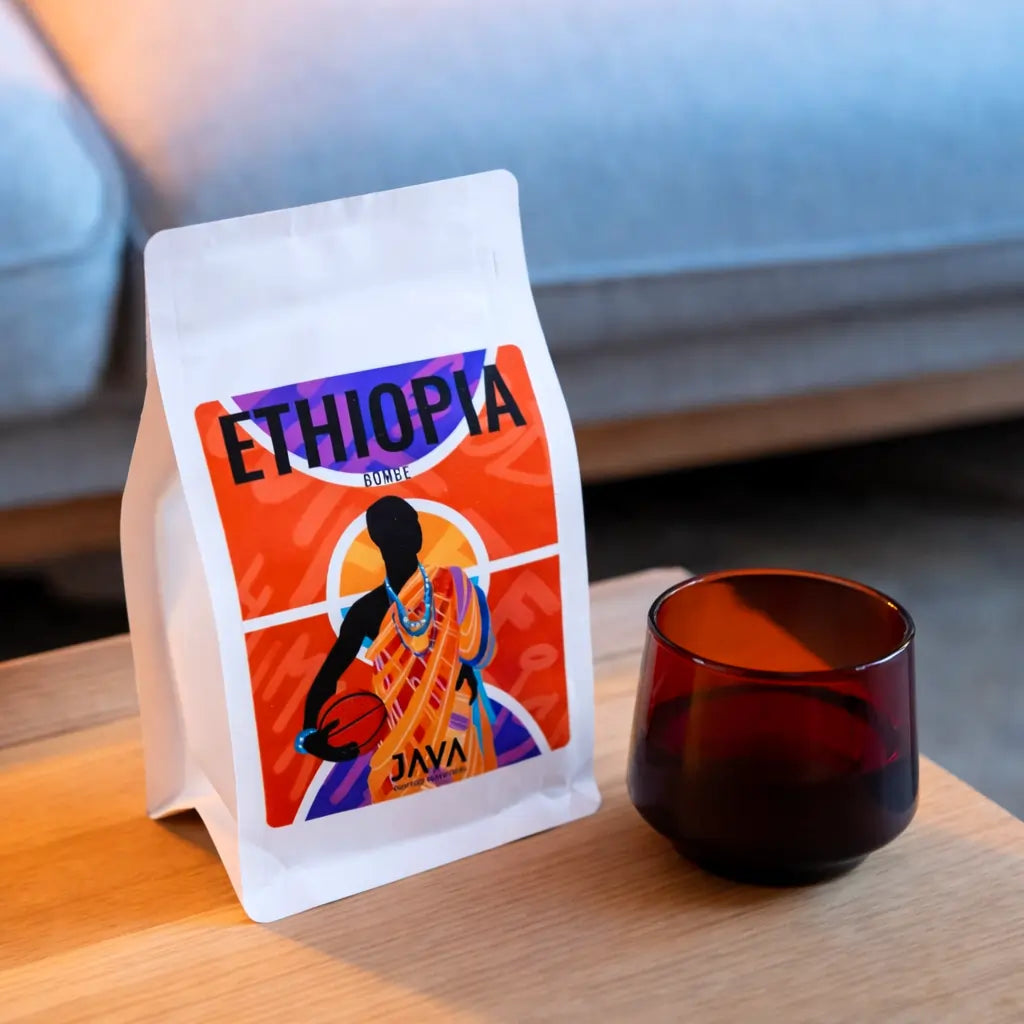 Kawa ziarnista Ethiopia Bombe JAVA Coffee Roasters – opakowanie kawy specialty na drewnianym stoliku obok szklanki, ujęcie lifestyle w ciepłym świetle