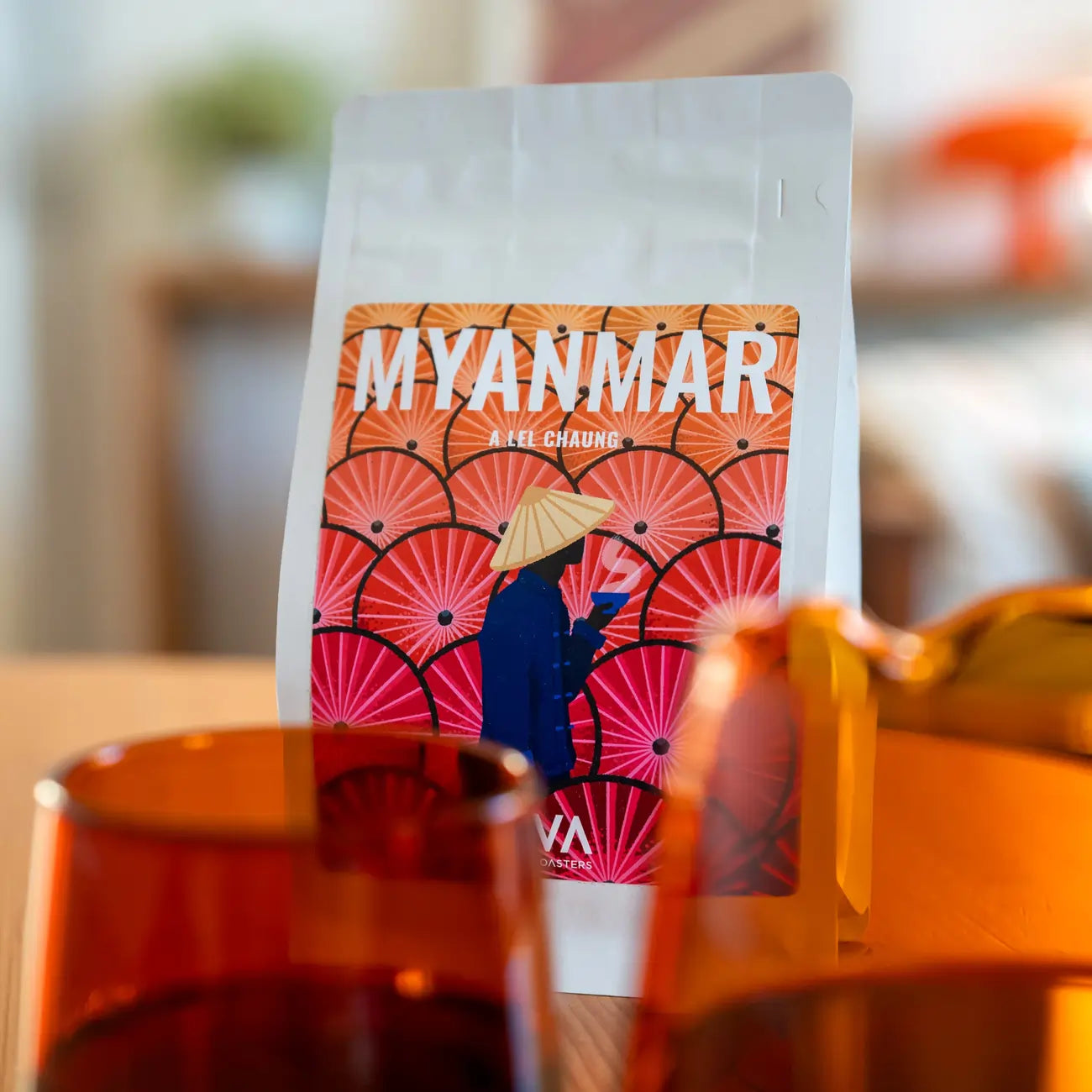 Opakowanie kawy ziarnistej Myanmar Ale Chaung od JAVA Coffee Roasters na drewnianym stole, w tle bursztynowe szkło