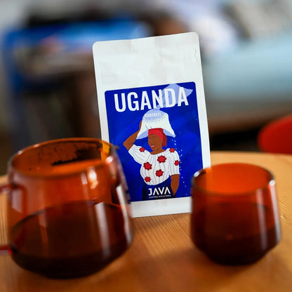 JAVA Coffee Roasters Uganda Rubirizi 250 g – kawa specialty 100% Arabica w ziarnach na drewnianym stole z serwerem i filiżanką we wnętrzu nowoczesnej kawiarni.