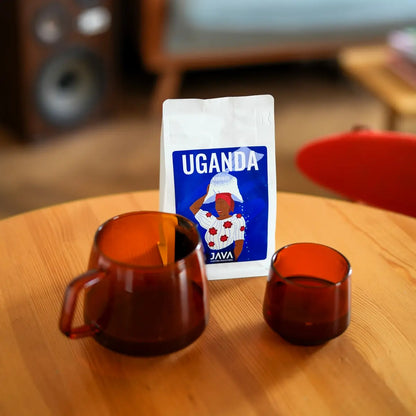 JAVA Coffee Roasters Uganda Rubirizi 250 g – kawa specialty 100% Arabica w ziarnach na drewnianym stole z serwerem i filiżanką do kawy we wnętrzu kawiarni.