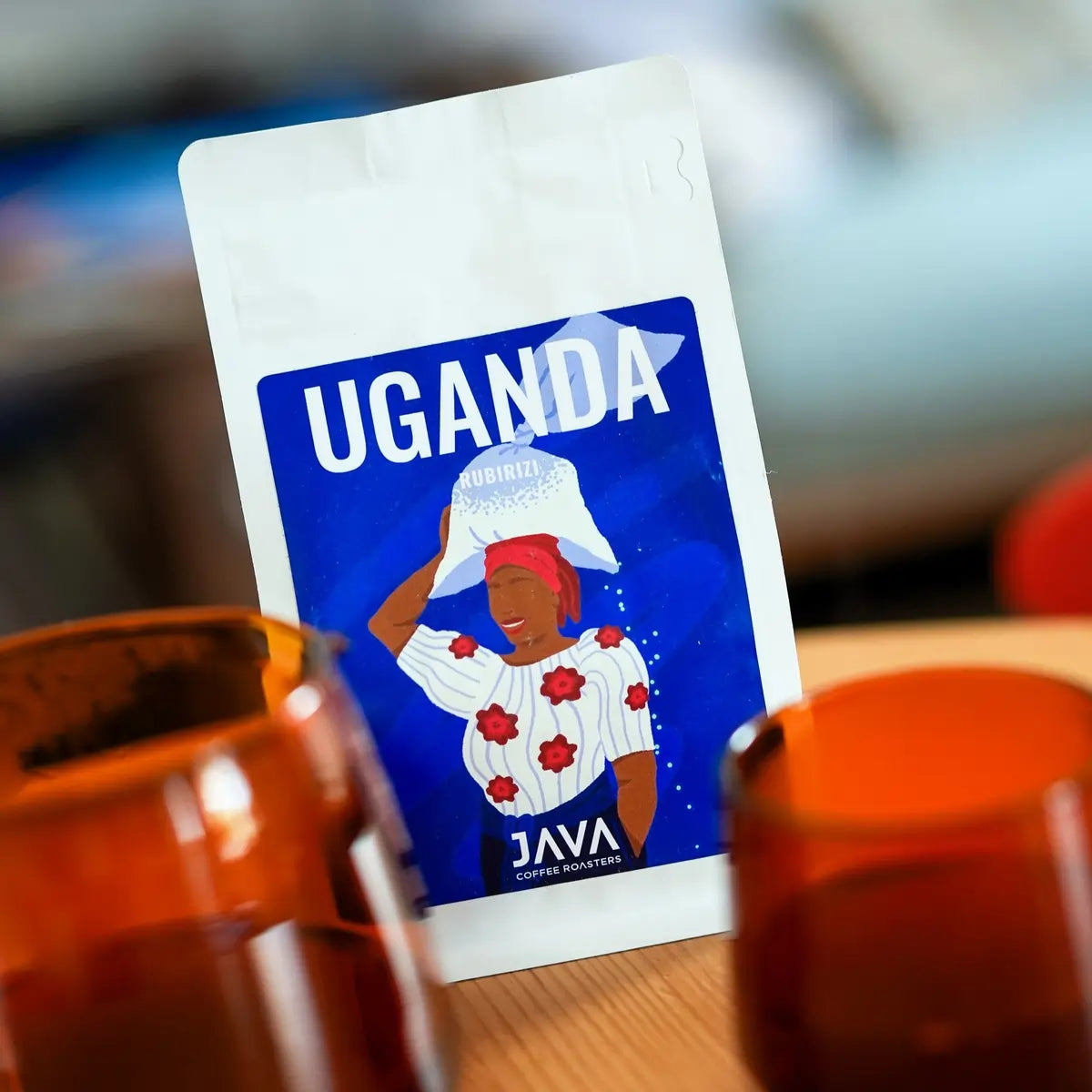 Zbliżenie na opakowanie kawy JAVA Coffee Roasters Uganda Rubirizi 250 g na drewnianym stole z filiżankami do kawy w nowoczesnej kawiarni – świeżo palona kawa specialty 100% Arabica.