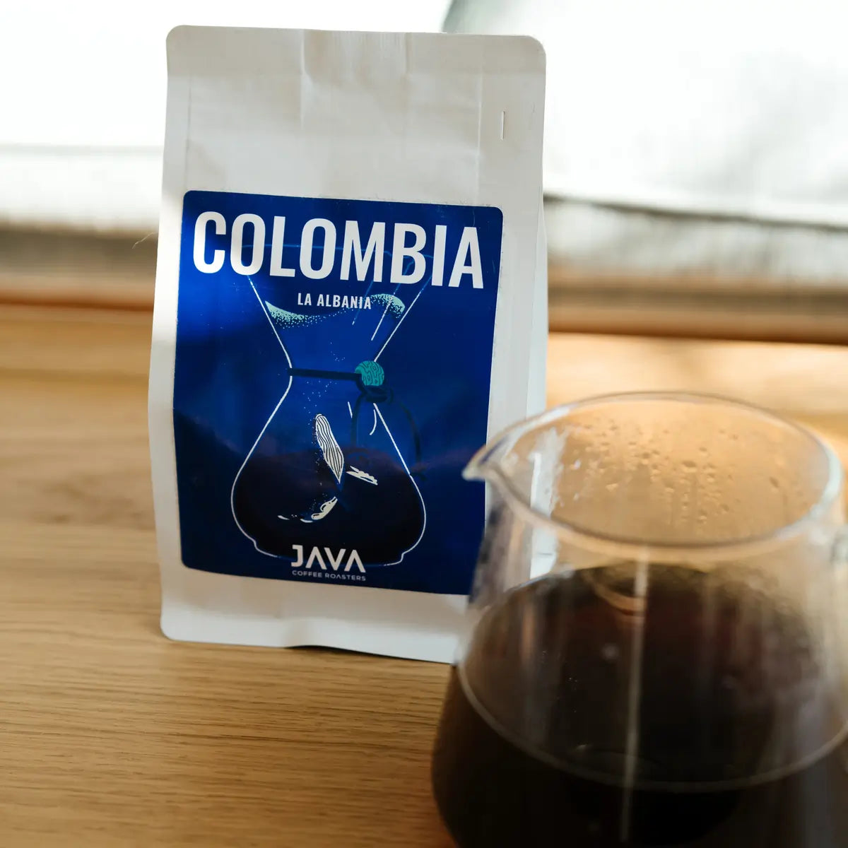 Kawa speciality Java Coffee Roasters Colombia La Albania w białym opakowaniu obok dzbanka z zaparzoną kawą na drewnianym stole