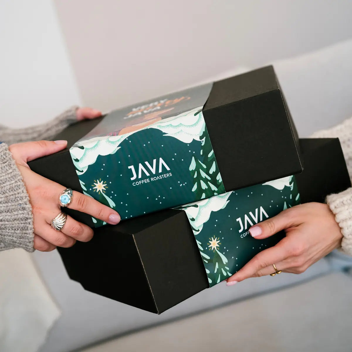 Świąteczne gift boxy JAVA Coffee Roasters trzymane w dłoniach — czarne pudełka z zimową grafiką Very Merry Java.