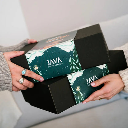 Świąteczne gift boxy JAVA Coffee Roasters trzymane w dłoniach — czarne pudełka z zimową grafiką Very Merry Java.