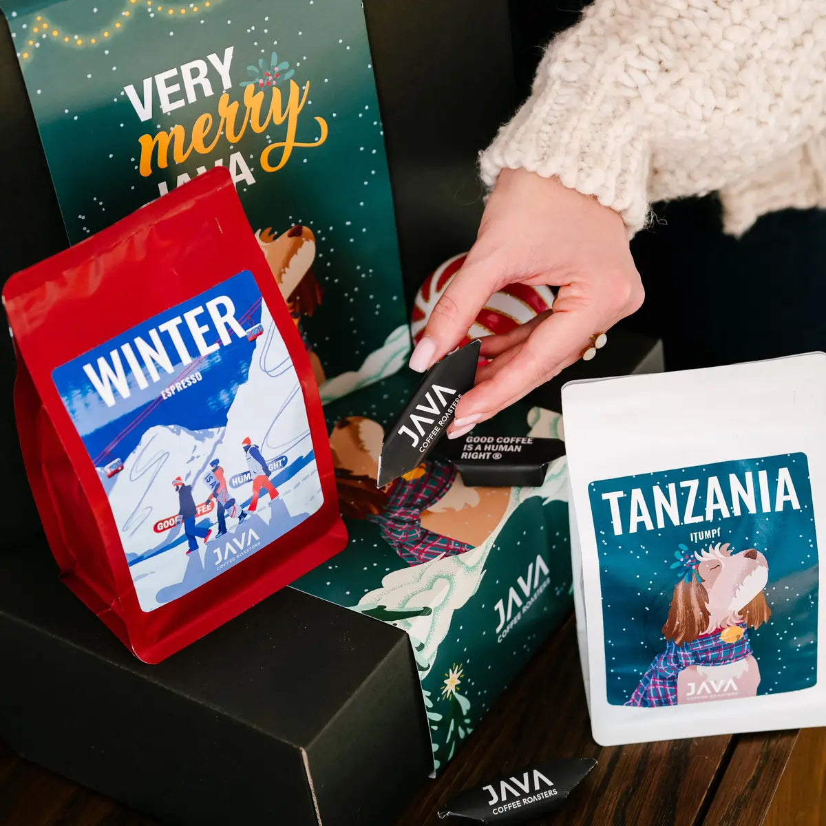 Zbliżenie na kawę Winter Espresso i Tanzania oraz świąteczne pudełko Very Merry Java, ustawione na czarnym boxie JAVA Coffee Roasters, z ozdobną bombką w tle.
