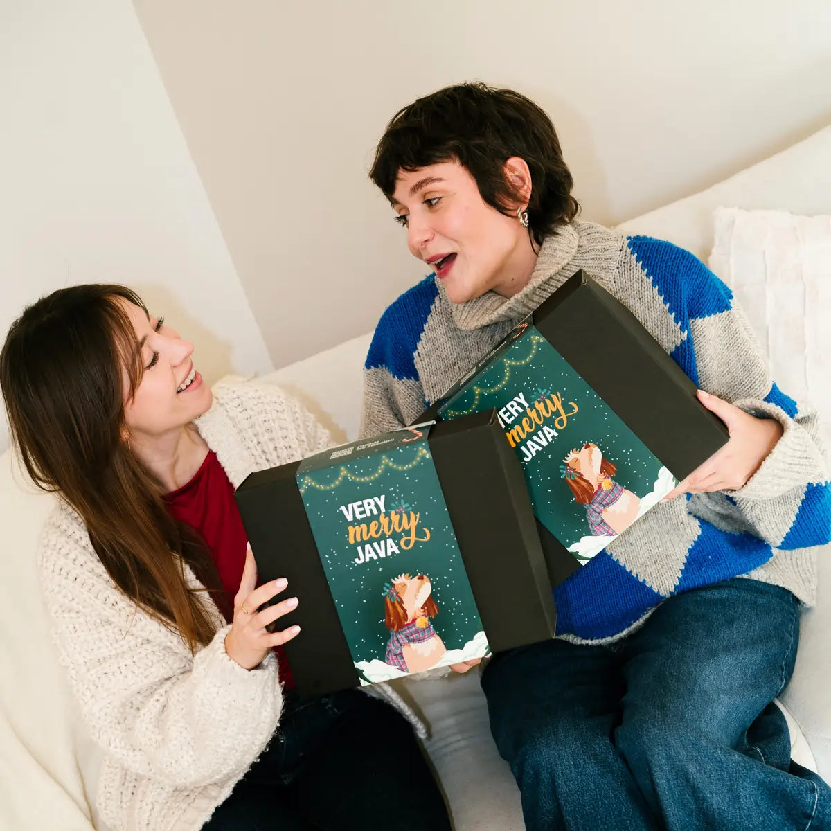 Dwie osoby trzymają świąteczne zestawy prezentowe Very Merry JAVA Coffee Roasters z zimową grafiką i psem na opakowaniu.