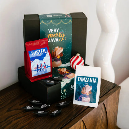 Zestaw świąteczny JAVA Coffee z kawami Winter Espresso i Tanzania Itumpi, czarnym pudełkiem Very Merry Java oraz cukierkami, ustawiony na drewnianej komodzie.