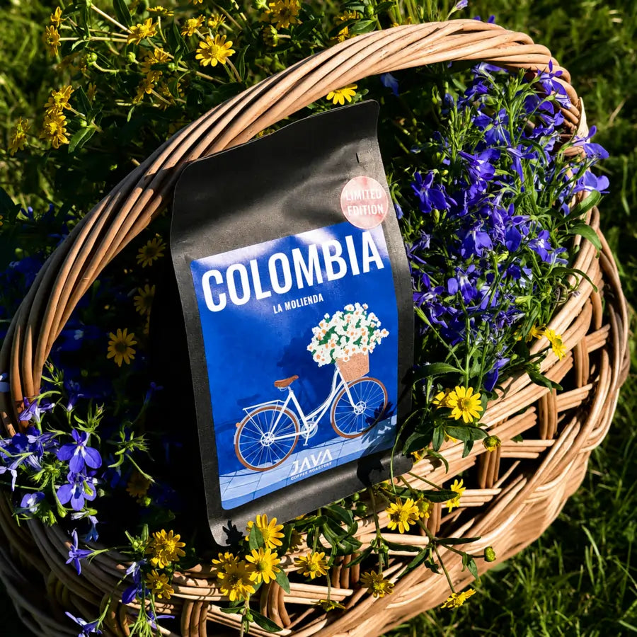 Opakowanie kawy ziarnistej Colombia La Molienda JAVA Coffee umieszczone w wiklinowym koszu wśród niebieskich i żółtych kwiatów.