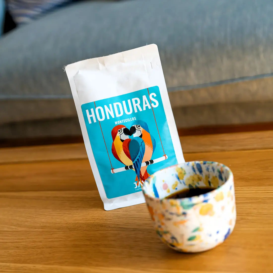 Kawa Java Honduras Montecillos w białym opakowaniu obok kolorowej filiżanki z zaparzoną kawą, ujęcie lifestyle na drewnianym stole