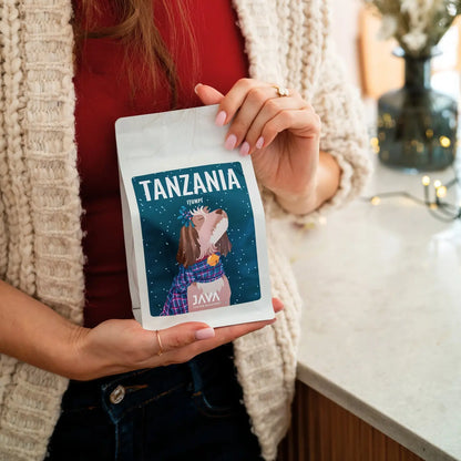 Kobieta trzyma opakowanie kawy JAVA Coffee Tanzania Itumpi, stojąc przy blacie w jasnym, przytulnym wnętrzu.