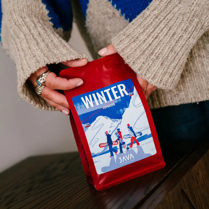 Osoba trzyma czerwone opakowanie kawy JAVA Coffee Winter Espresso, ustawiając je na stole w domowym wnętrzu